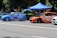 Two modified cars are parked on the side of a road under a blue canopy. The blue car features prominent branding with 'Grill Market' on the side, while the orange car has its hood open, indicating it might be undergoing maintenance or inspection. Both vehicles display various sponsor logos and decals and have racing numbers. The background shows a park-like setting with trees and a few people.