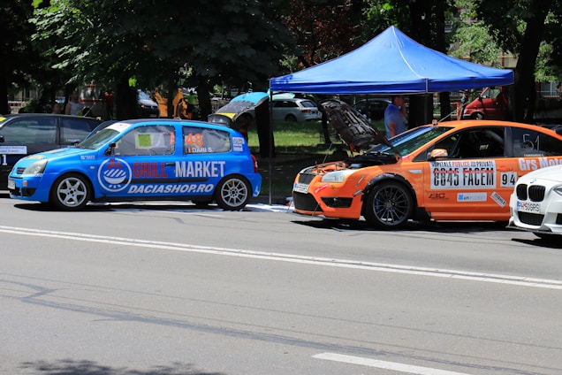 Two modified cars are parked on the side of a road under a blue canopy. The blue car features prominent branding with 'Grill Market' on the side, while the orange car has its hood open, indicating it might be undergoing maintenance or inspection. Both vehicles display various sponsor logos and decals and have racing numbers. The background shows a park-like setting with trees and a few people.