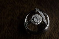 A close-up view of a diamond engagement ring and wedding band resting against a textured dark background. The rings are intricately designed, featuring a large central diamond surrounded by smaller stones, all set in a shiny metal band.