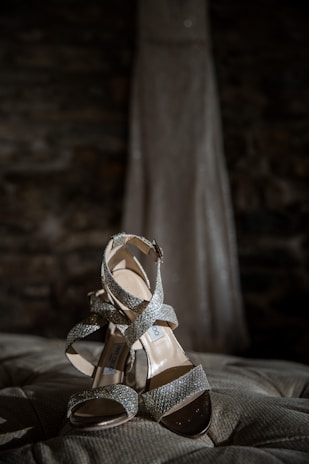A pair of sparkling, high-heeled sandals with crisscross straps rests elegantly on a tufted surface. In the background, the blurred silhouette of a long gown can be seen.