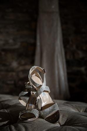 A pair of sparkling, high-heeled sandals with crisscross straps rests elegantly on a tufted surface. In the background, the blurred silhouette of a long gown can be seen.