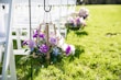 A decorative arrangement featuring purple and pink flowers with greenery is hung on a metal stand. The setting includes white wooden chairs lined up on a lush green lawn, suggesting an outdoor event or ceremony.
