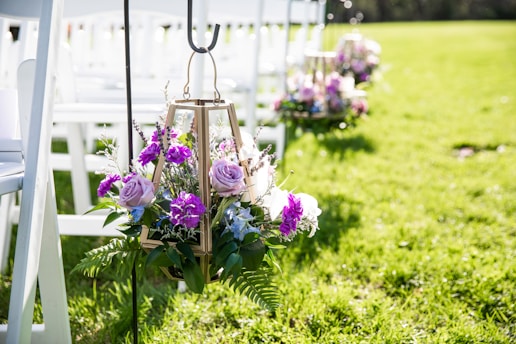 A decorative arrangement featuring purple and pink flowers with greenery is hung on a metal stand. The setting includes white wooden chairs lined up on a lush green lawn, suggesting an outdoor event or ceremony.