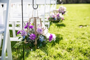A decorative arrangement featuring purple and pink flowers with greenery is hung on a metal stand. The setting includes white wooden chairs lined up on a lush green lawn, suggesting an outdoor event or ceremony.