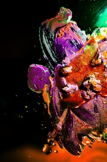 Vibrant strokes of paint blend together in an abstract composition featuring vivid purples, oranges, and greens. Thick applications of paint create texture and depth, with interspersed splashes and droplets adding dynamic movement.
