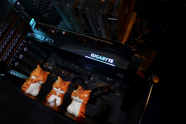 Inside a computer case, a large graphics card labeled 'GEFORCE RTX' and 'GIGABYTE' is mounted, surrounded by cooling components and cables. In front of the card are three small, orange and white cat figurines with different poses, adding a playful element to the technical setup.