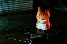 A small figurine of a cartoon-like orange cat with stripes sits on a dark surface. The cat appears to be meditating or sitting in a relaxed pose with its eyes closed. Nearby, there is a bundle of black computer cables with labels and a circuit board visible in the foreground. The lighting is dim with a warm glow on the cat and cooler tones on the background.