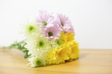 A bouquet of flowers featuring a combination of soft pink, white, and bright yellow blooms, arranged atop a light wooden surface. The flowers have a fresh, vibrant appearance with lush, textured petals and leafy green centers.