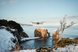 A drone hovers over a winter coastal landscape. Snow-covered cliffs and rocky formations rise from the shallow waters, with a distant view of the sea and a partly cloudy sky. Leafless branches of a tree are visible to the right.