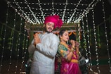 A couple dressed in traditional Indian attire are posing back-to-back under a canopy of white string lights. The man is wearing a white sherwani with a bright pink turban, while the woman is dressed in a pink and green saree with intricate jewelry and henna on her hands.