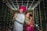 A couple dressed in traditional Indian attire are posing back-to-back under a canopy of white string lights. The man is wearing a white sherwani with a bright pink turban, while the woman is dressed in a pink and green saree with intricate jewelry and henna on her hands.