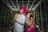A couple dressed in traditional Indian attire are posing back-to-back under a canopy of white string lights. The man is wearing a white sherwani with a bright pink turban, while the woman is dressed in a pink and green saree with intricate jewelry and henna on her hands.