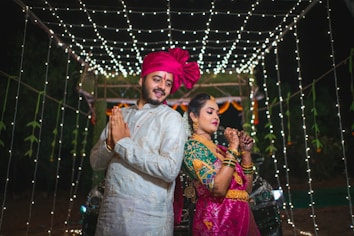 A couple dressed in traditional Indian attire are posing back-to-back under a canopy of white string lights. The man is wearing a white sherwani with a bright pink turban, while the woman is dressed in a pink and green saree with intricate jewelry and henna on her hands.