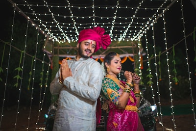A couple dressed in traditional Indian attire are posing back-to-back under a canopy of white string lights. The man is wearing a white sherwani with a bright pink turban, while the woman is dressed in a pink and green saree with intricate jewelry and henna on her hands.