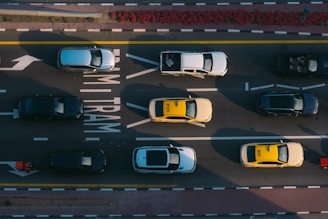 Aerial view of cars and taxis moving on a multi-lane road marked with 'TRAM' lanes. Vehicles are arranged in a pattern with taxis in bright yellow and other cars in darker shades. The lanes are bordered by solid white and yellow lines.