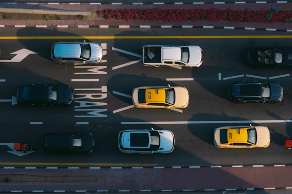 Aerial view of cars and taxis moving on a multi-lane road marked with 'TRAM' lanes. Vehicles are arranged in a pattern with taxis in bright yellow and other cars in darker shades. The lanes are bordered by solid white and yellow lines.