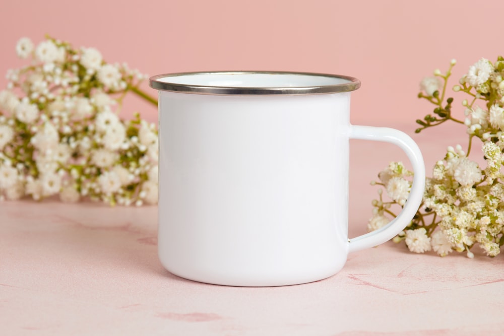 a white mug with a silver rim sitting next to a bunch of white flowers