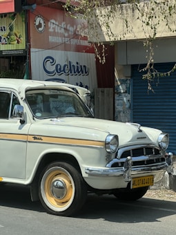 A vintage white car with yellow accents is parked in front of a closed store with signage displaying Cochin fruits and a supermarket ad. The car has a classic design with prominent round headlights and a chrome grille. On the car's license plate, the registration number is visible. There are some leaves and branches hanging from above.