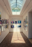 A well-lit hallway with a glass ceiling displaying several framed posters on both walls. The posters feature various artistic designs and are part of an exhibition. The center wall displays the name 'Yves Saint Laurent' above the posters.