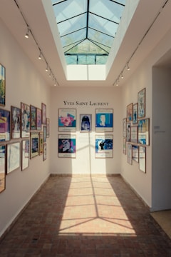 A well-lit hallway with a glass ceiling displaying several framed posters on both walls. The posters feature various artistic designs and are part of an exhibition. The center wall displays the name 'Yves Saint Laurent' above the posters.