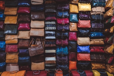 A variety of colorful bags are densely stacked against a wall, forming an organized display. Each bag comes in different shapes and textures, showcasing a wide range of materials such as leather and fabric. The bags are arranged in columns and rows, creating a visually striking pattern.