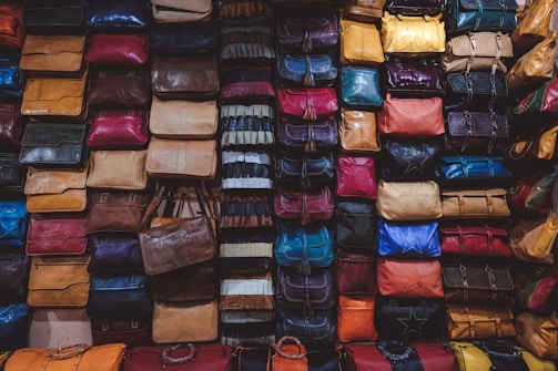 A variety of colorful bags are densely stacked against a wall, forming an organized display. Each bag comes in different shapes and textures, showcasing a wide range of materials such as leather and fabric. The bags are arranged in columns and rows, creating a visually striking pattern.