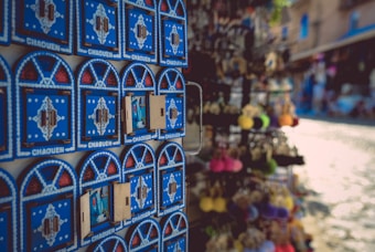 A display of vibrant blue fridge magnets with intricate designs and the word 'CHAQUEN' written on them. The background shows a blurred view of a sunlit street market with various colorful items on display.