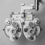 A mechanical phoropter device used in optometry, featuring multiple dials, lenses, and adjustment knobs. The device is predominantly metallic and white, with intricate details and markings for precise adjustments. It is used to measure the refractive error and determine eyeglass prescriptions.