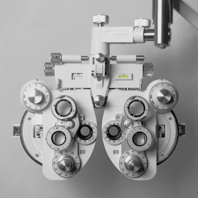 A mechanical phoropter device used in optometry, featuring multiple dials, lenses, and adjustment knobs. The device is predominantly metallic and white, with intricate details and markings for precise adjustments. It is used to measure the refractive error and determine eyeglass prescriptions.