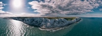 A panoramic view of the white cliffs bordering a calm, expansive sea under a partly cloudy sky with the sun shining brightly. The landscape includes lush greenery atop the cliffs, stretching along the coastline.