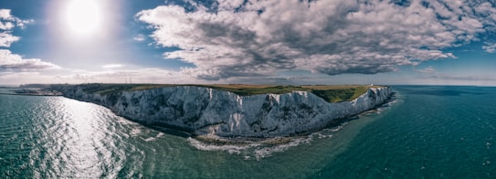 A panoramic view of the white cliffs bordering a calm, expansive sea under a partly cloudy sky with the sun shining brightly. The landscape includes lush greenery atop the cliffs, stretching along the coastline.