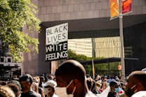 A crowd of people gather at a protest with many wearing face masks. A prominent sign in the middle reads 'Black Lives Over White Feelings'. The scene is set in an urban environment with buildings and a large tree.