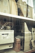 Several outdated electronic devices, including vintage computer monitors and printers, are stacked on shelves behind a glass window. The equipment is mostly beige with signs of wear and aging.