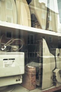 Several outdated electronic devices, including vintage computer monitors and printers, are stacked on shelves behind a glass window. The equipment is mostly beige with signs of wear and aging.
