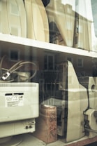 Several outdated electronic devices, including vintage computer monitors and printers, are stacked on shelves behind a glass window. The equipment is mostly beige with signs of wear and aging.
