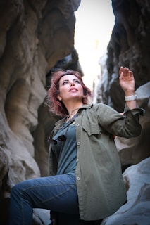 A person with reddish-brown hair wearing a green jacket and blue pants stands in a rocky, narrow canyon. The person is looking up with one arm raised, and they have sunglasses hanging on their chest.