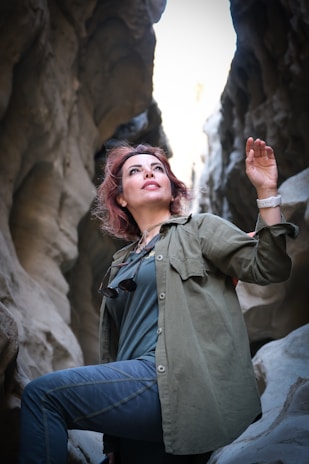 A person with reddish-brown hair wearing a green jacket and blue pants stands in a rocky, narrow canyon. The person is looking up with one arm raised, and they have sunglasses hanging on their chest.