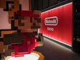 A pixelated sculpture, reminiscent of classic video game graphics, depicts a character with a red hat and shirt. It is placed inside a modern space with a large, illuminated sign in the background bearing the Nintendo logo and the word Tokyo.