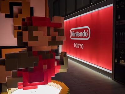 A pixelated sculpture, reminiscent of classic video game graphics, depicts a character with a red hat and shirt. It is placed inside a modern space with a large, illuminated sign in the background bearing the Nintendo logo and the word Tokyo.
