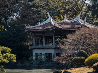 A traditional Asian pavilion with a curved roof is surrounded by dense greenery and trees. The structure stands elevated on pillars over a rock garden. The atmosphere suggests tranquility and harmony with nature.