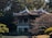 A traditional Asian pavilion with a curved roof is surrounded by dense greenery and trees. The structure stands elevated on pillars over a rock garden. The atmosphere suggests tranquility and harmony with nature.