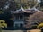 A traditional Asian pavilion with a curved roof is surrounded by dense greenery and trees. The structure stands elevated on pillars over a rock garden. The atmosphere suggests tranquility and harmony with nature.