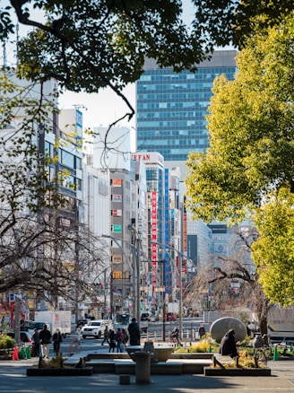 An urban scene depicting a busy street in a city setting, featuring tall buildings with advertisements and signages in various colors. The foreground shows people walking and sitting in a park area, surrounded by trees with both green and bare branches. A mix of modern architecture and urban nature creates a dynamic blend.