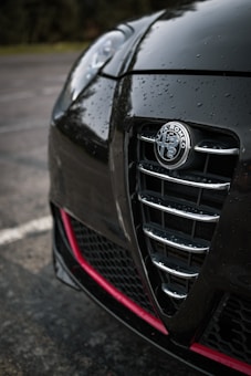 Close-up of a black car featuring rain droplets on its surface. The focus is on the front grille with a prominent Alfa Romeo logo. The surrounding background is slightly blurred, possibly an outdoor parking lot.