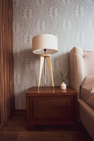 A cozy bedroom corner featuring a wooden bedside table with a tan-colored lampshade on a wooden tripod stand. Beside it, a small white vase with green foliage rests on the table. The background showcases a textured wallpaper with a leaf pattern in gray tones, while the floor is made of wooden panels. The edge of a cushioned bed with a neutral fabric is visible on the right.