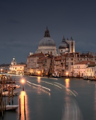 A scenic view of a canal in Venice, enveloped by historic buildings with ornate architecture. The image captures the iconic domes and bell towers against a dusky sky, while lights along the canal create a serene reflection on the water.