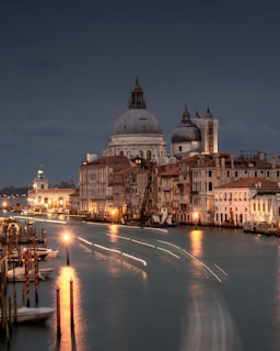 A scenic view of a canal in Venice, enveloped by historic buildings with ornate architecture. The image captures the iconic domes and bell towers against a dusky sky, while lights along the canal create a serene reflection on the water.