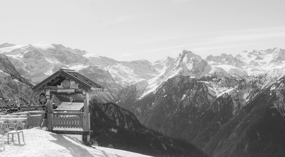 A snowy, mountainous landscape with a wooden structure featuring circular signs and an overhang. The backdrop consists of rugged mountain peaks covered in snow, under a clear sky. In the foreground, there are empty tables and chairs on a snow-covered terrace.