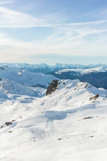 A winter landscape featuring snow-covered mountains under a clear blue sky. The pristine white snow blankets the peaks and slopes, creating a serene and peaceful atmosphere. Ski tracks are visible on the slopes, suggesting recreational activity.