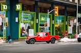 A bright red pickup truck is stopped at a crosswalk in front of a building featuring large green-framed windows. The building is branded with the TD logo and has posters with investment-themed messages. Snow is seen on the ground, and a person carrying a blue shopping bag is walking across the crosswalk.